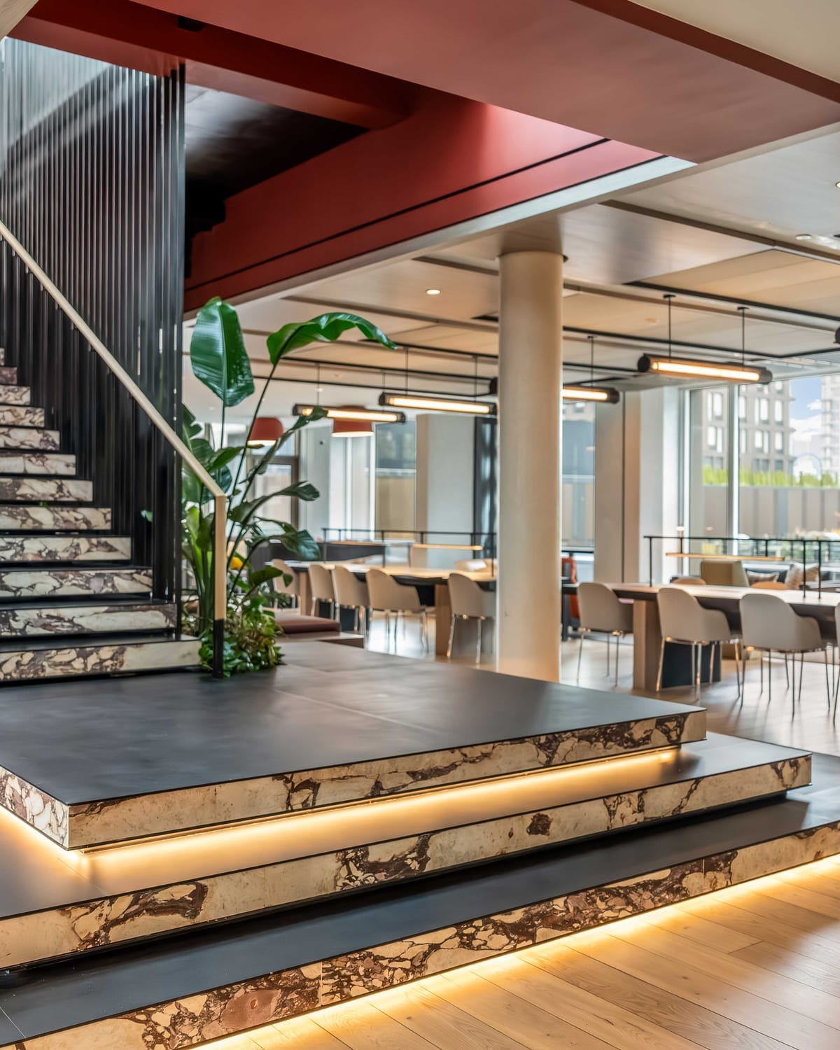 Interior staircase with illuminated steps, dark railing, plants, columns, and seating area beyond