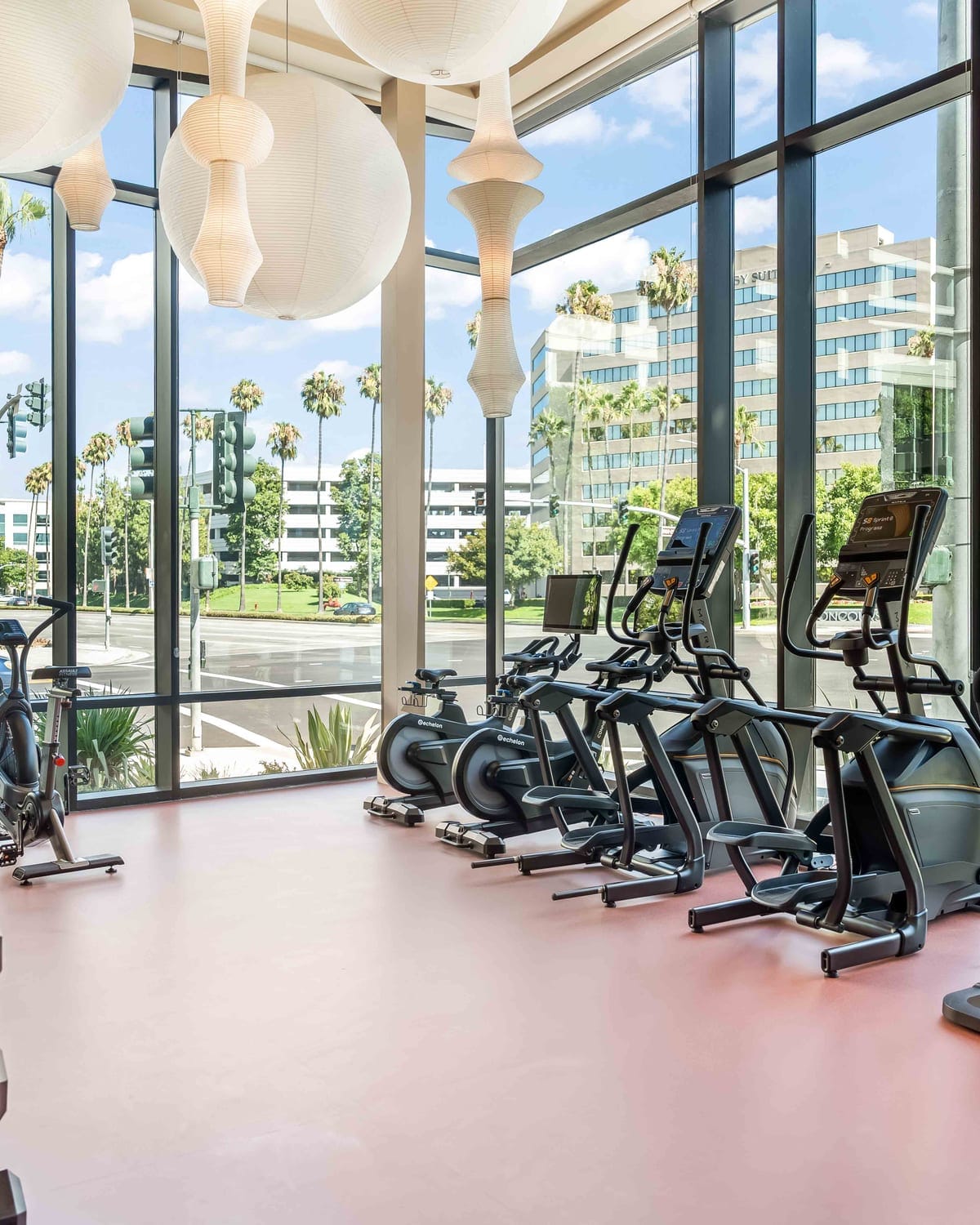 Cardio machines beside floor-to-ceiling windows beneath large hanging globe-shaped ceiling lights