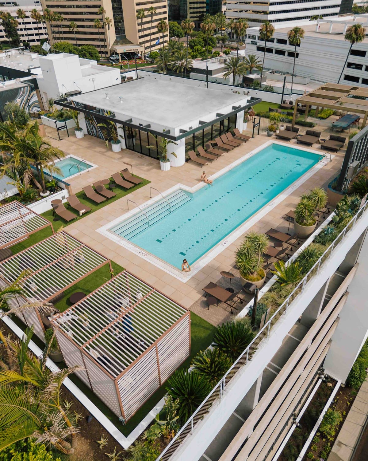 Elevated view of rectangular outdoor pool surrounded by loungers, pergolas, planters, and nearby buildings