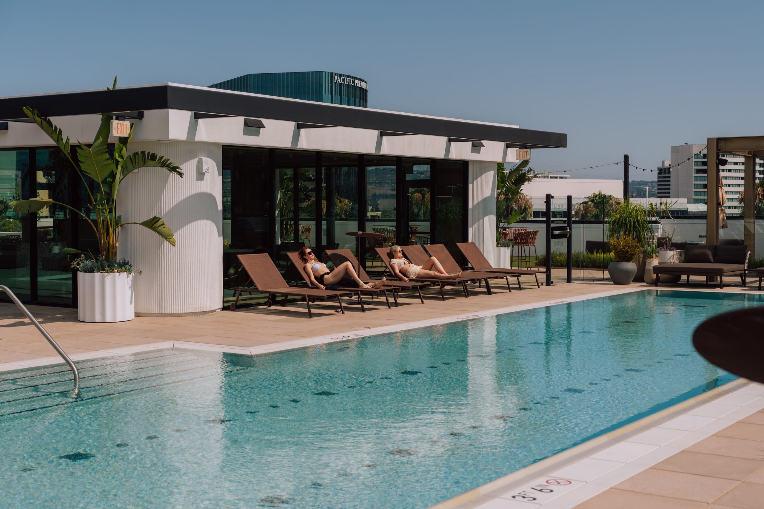 Outdoor pool beside loungers and low poolside building with plants under clear sky