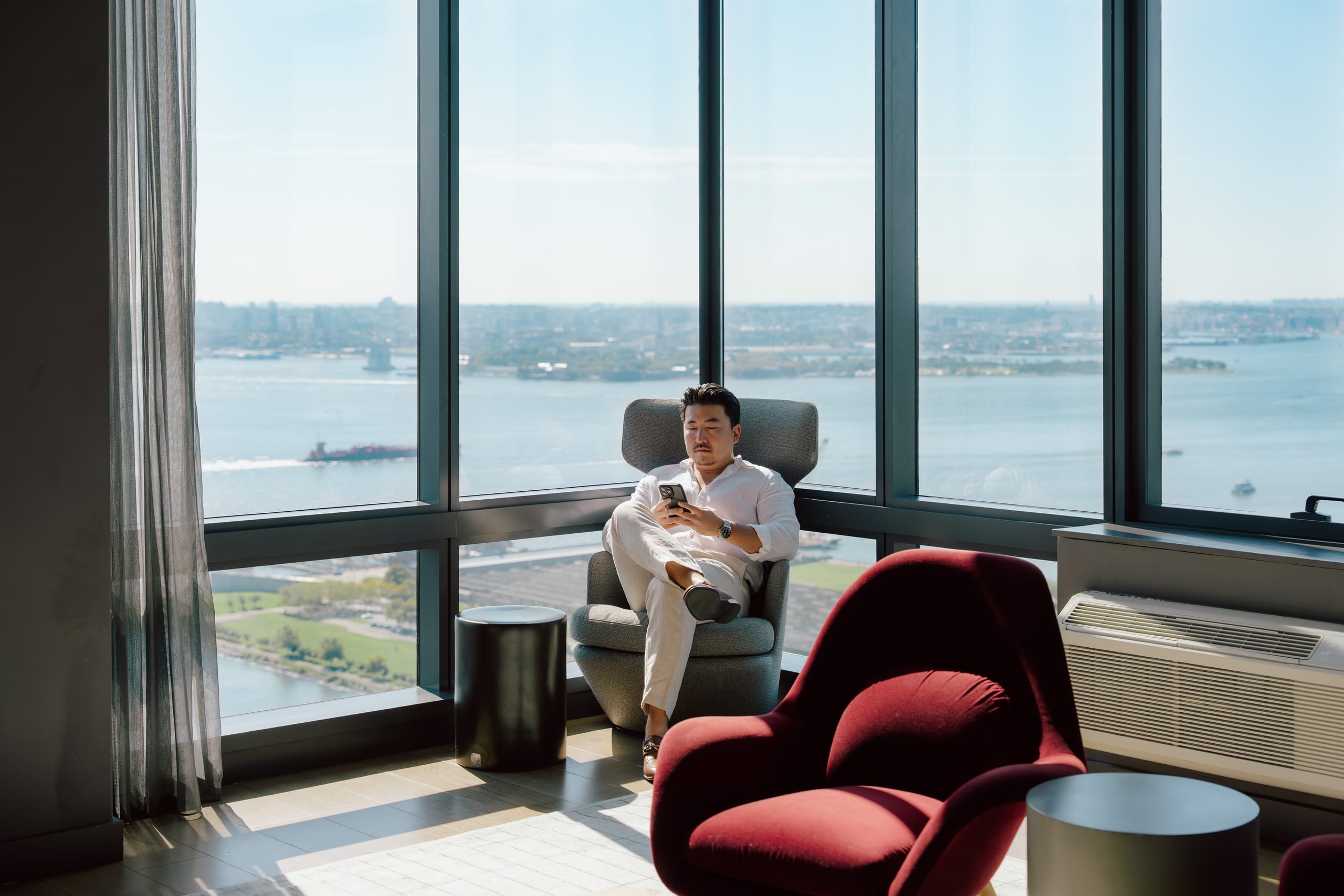 Person seated in armchair beside broad windows overlooking water and distant buildings