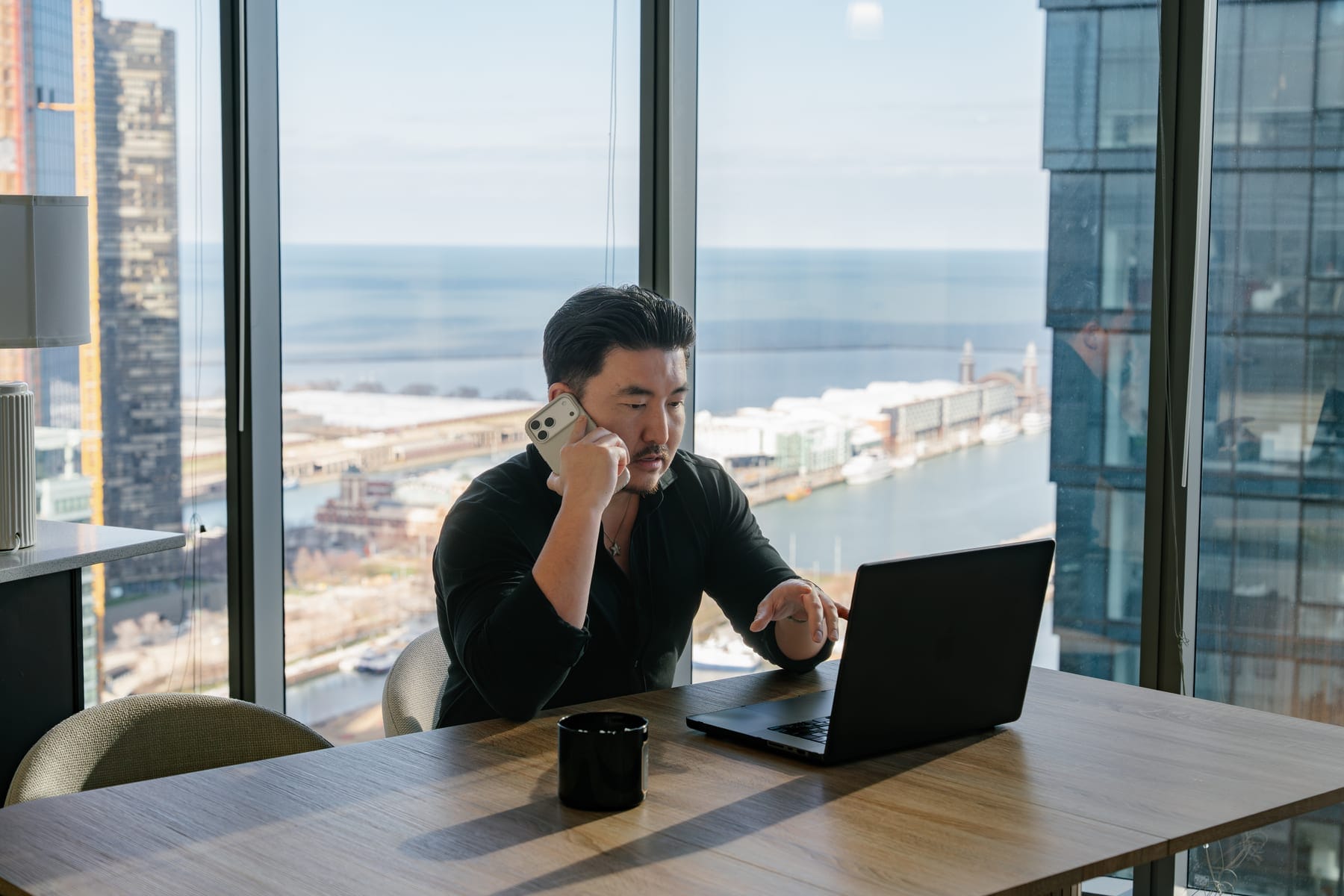 Person using laptop at dining table beside tall windows overlooking water and buildings