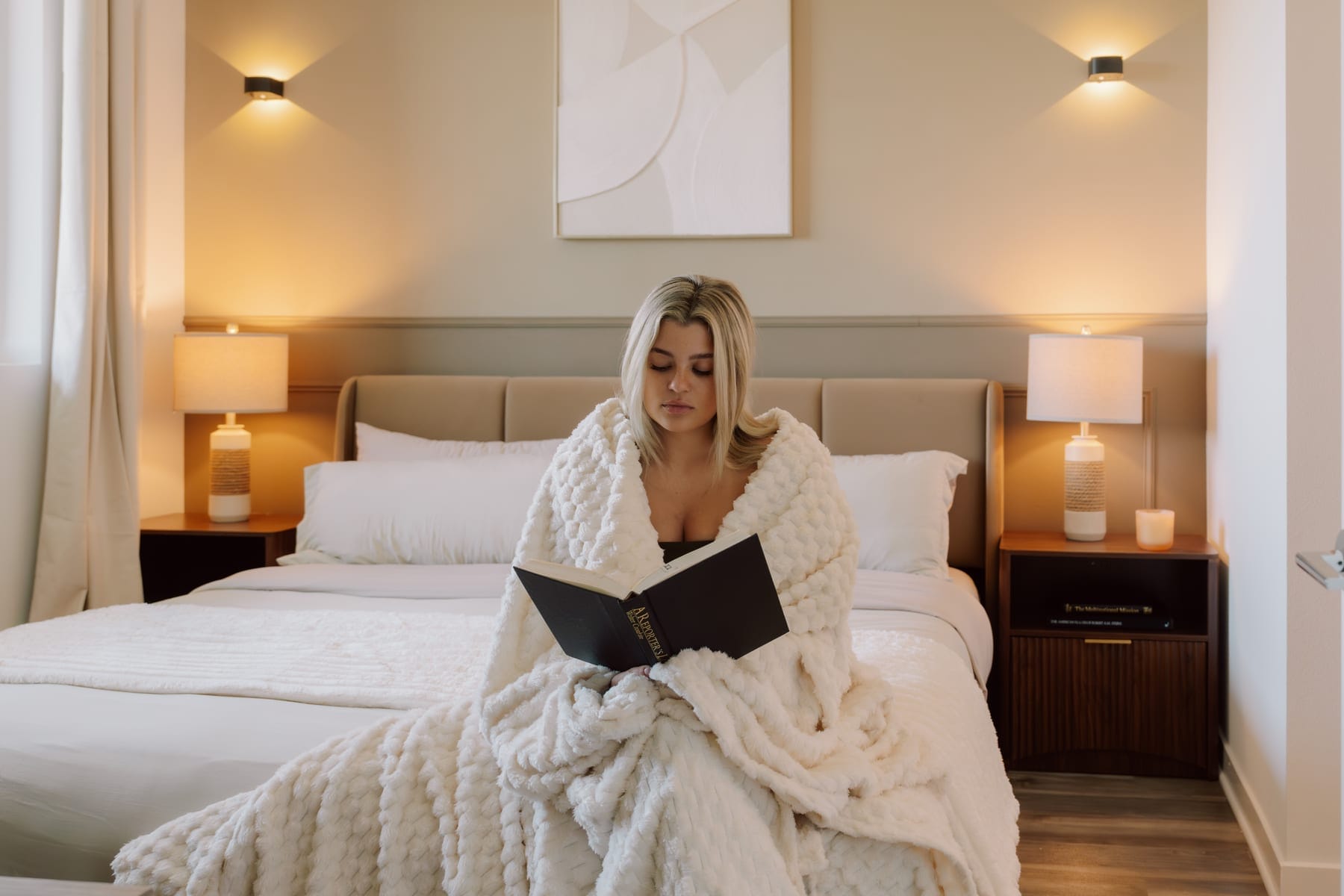 Woman wrapped in a white blanket reading a book while seated on a bed