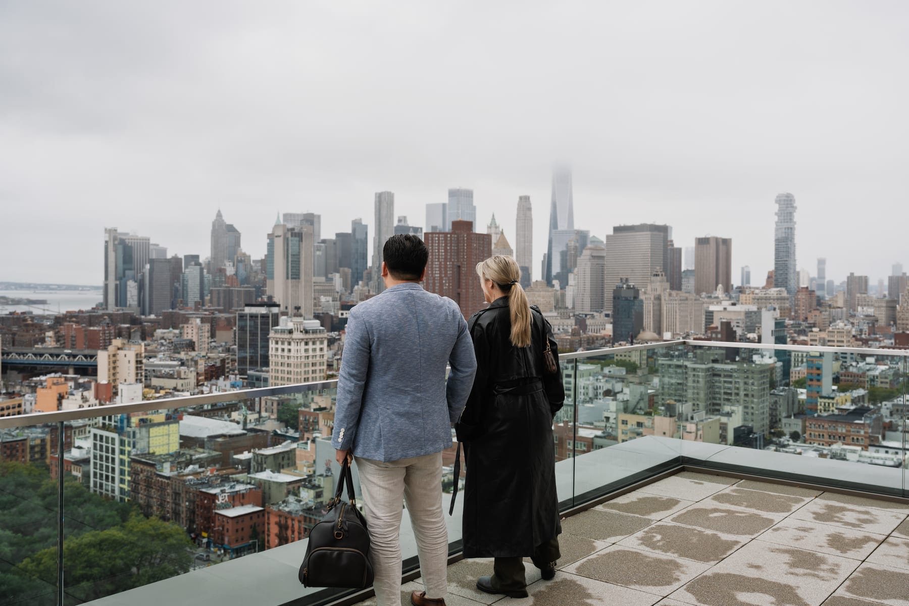 Two people standing at a rooftop glass railing looking across a distant city skyline