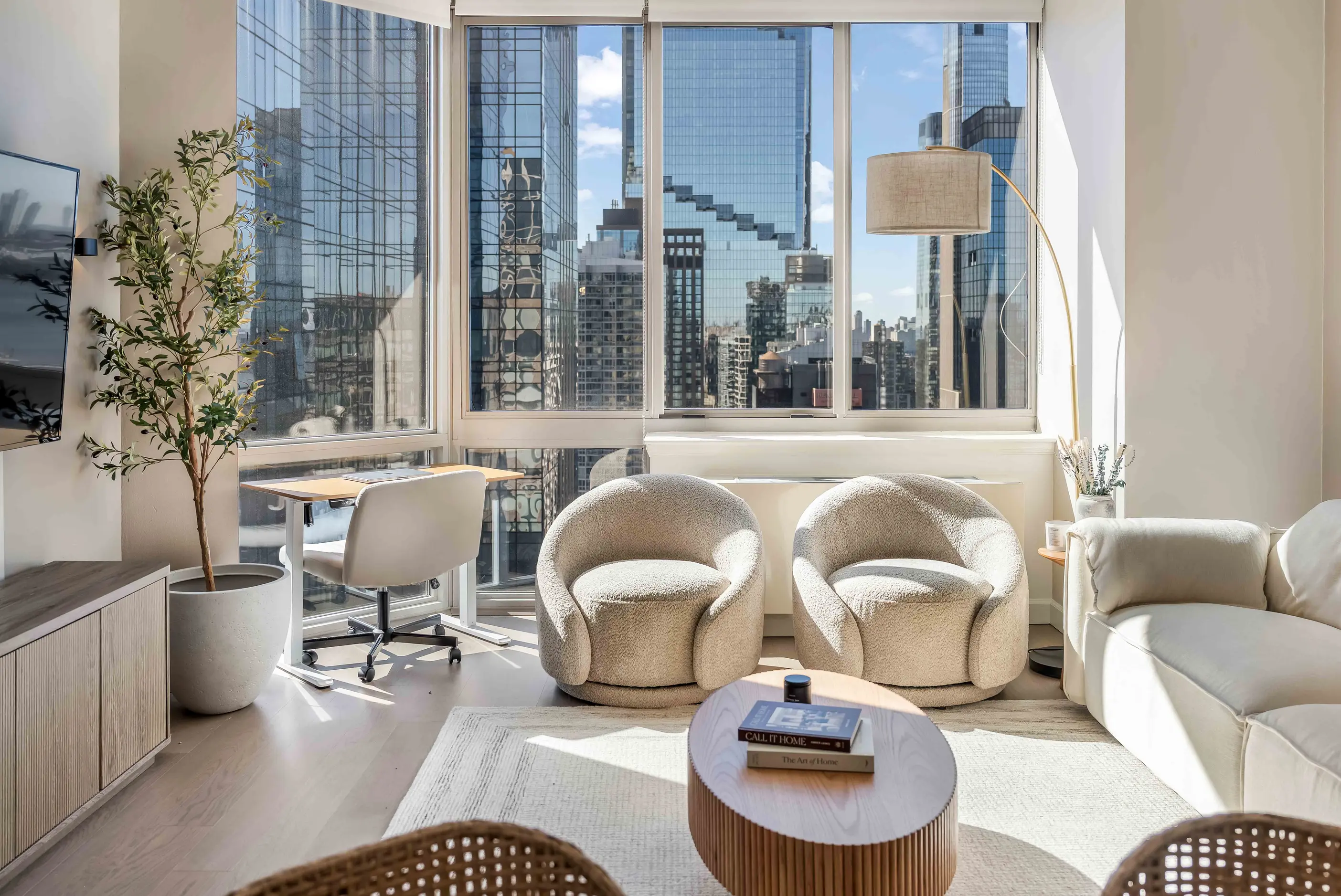 Living room with two rounded chairs, table, sofa, and desk beside tall city-facing windows at The Regent Highrise in New York City