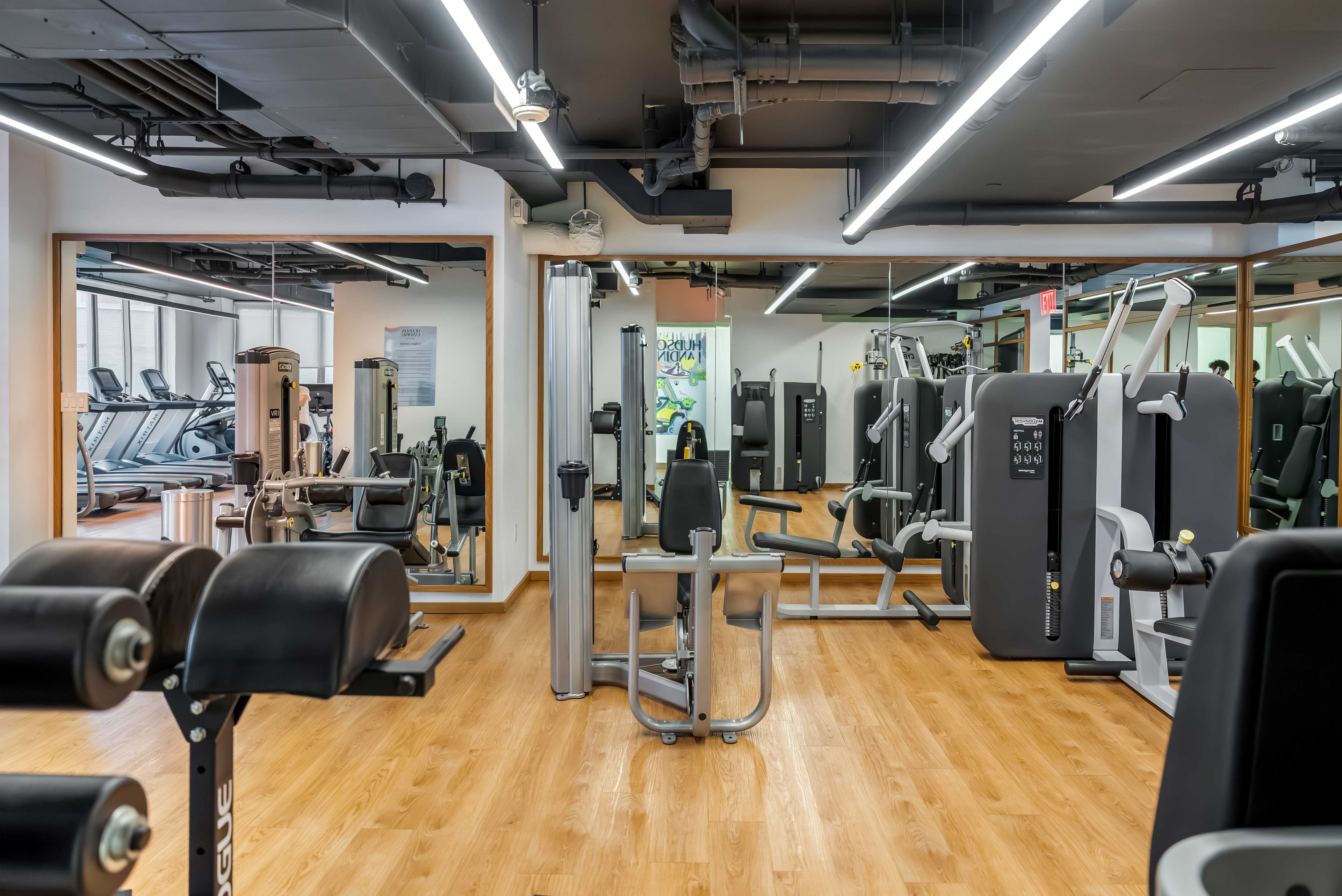 Gym with strength equipment beneath exposed ceiling and long bright overhead lights at Hudson Landing in New York City