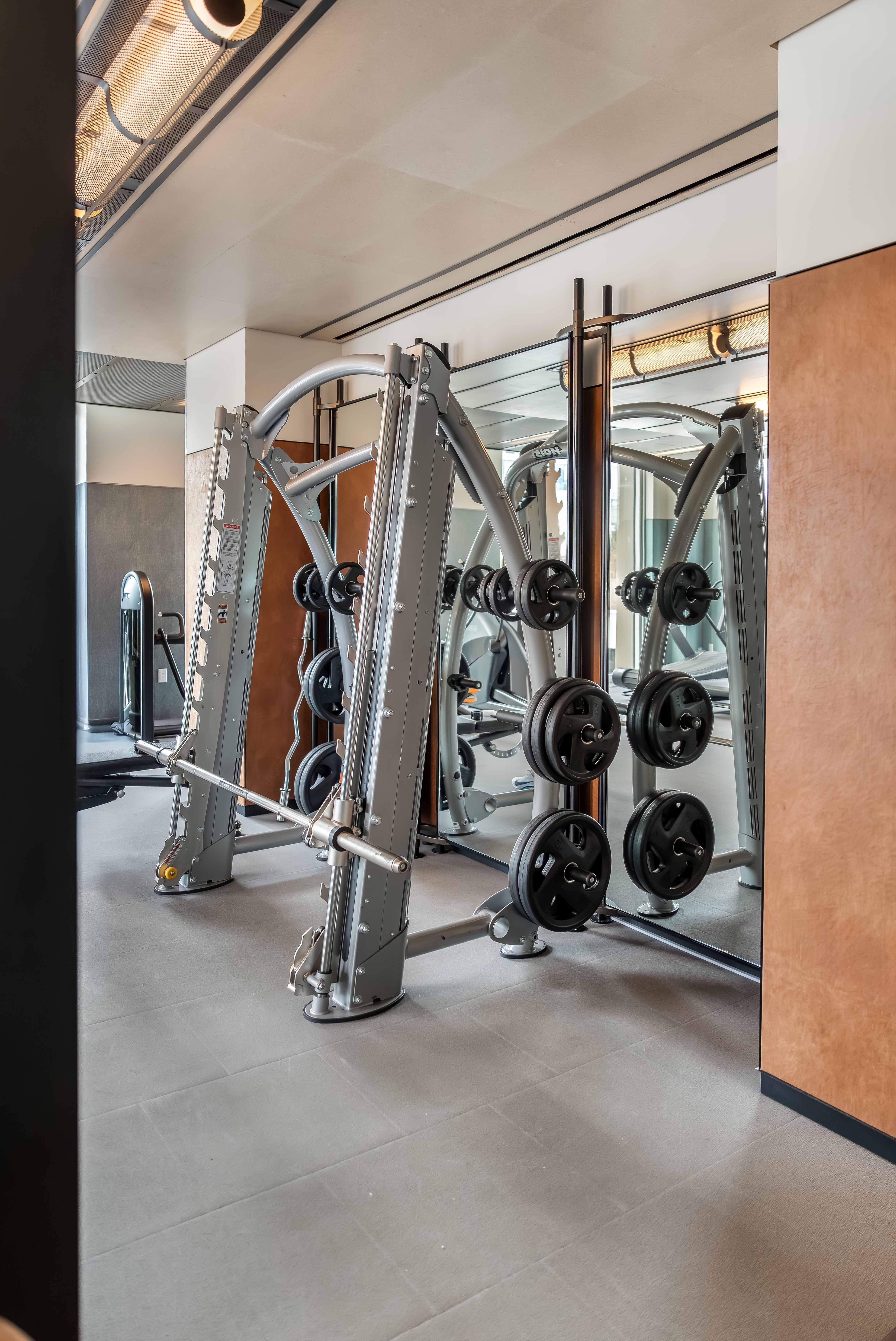 Gym strength machines with weight plates beside mirrored wall and exposed ceiling beams at The Suffolk in New York City