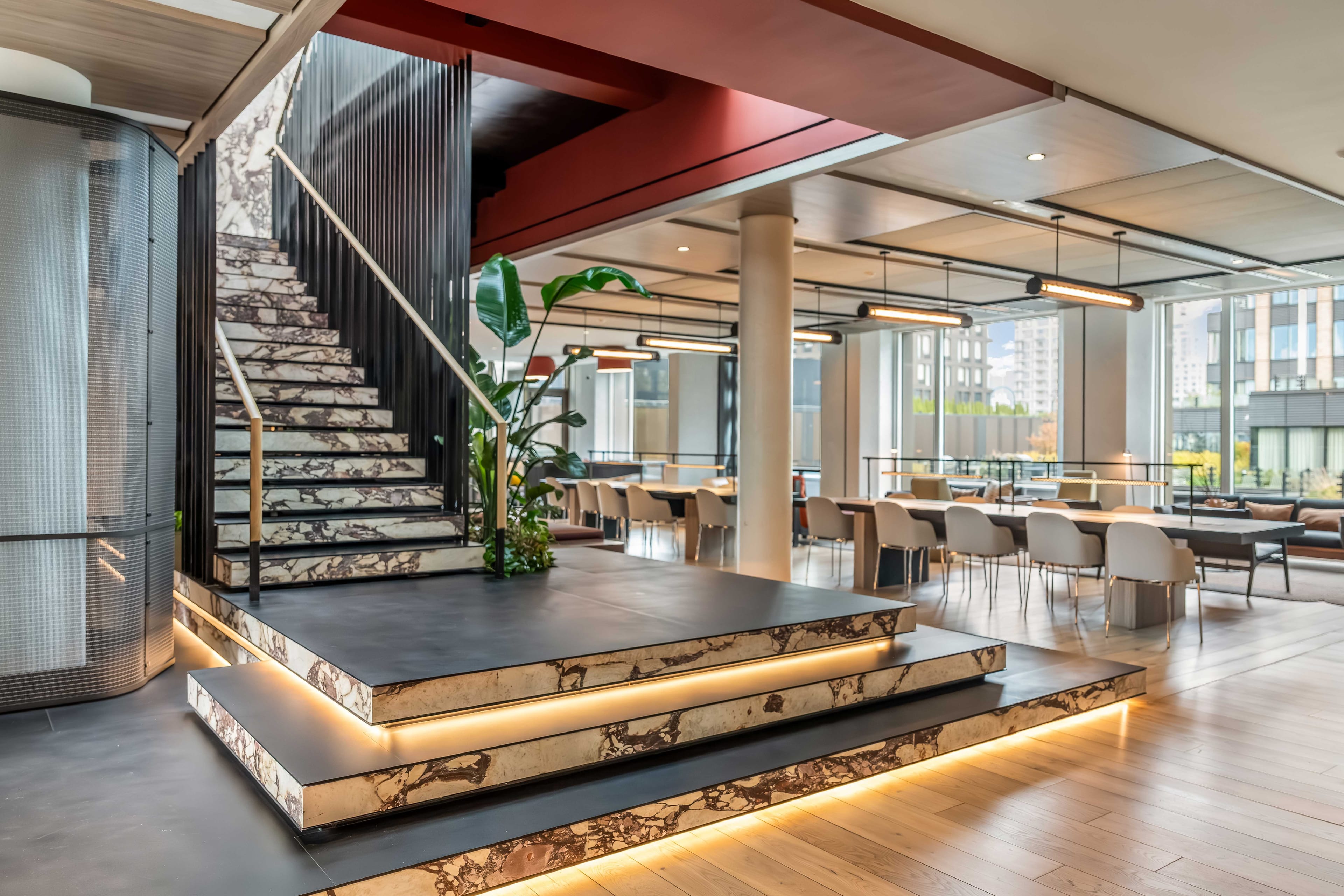 Shared lounge with open staircase, illuminated lower steps, dining tables, and broad windows at The Suffolk in New York City
