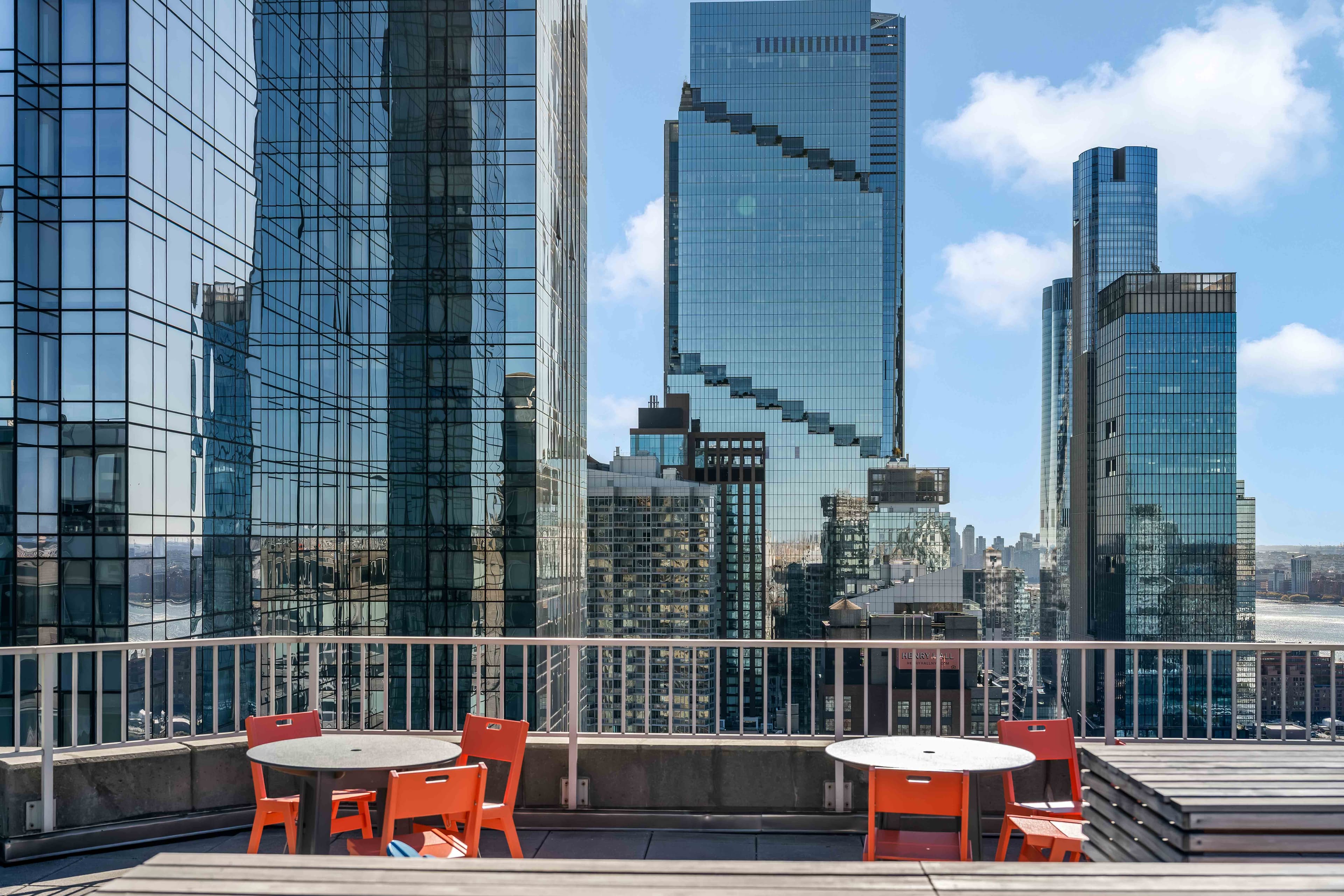 Rooftop terrace orange tables and chairs beneath glass towers and blue sky at Hudson Landing in New York City