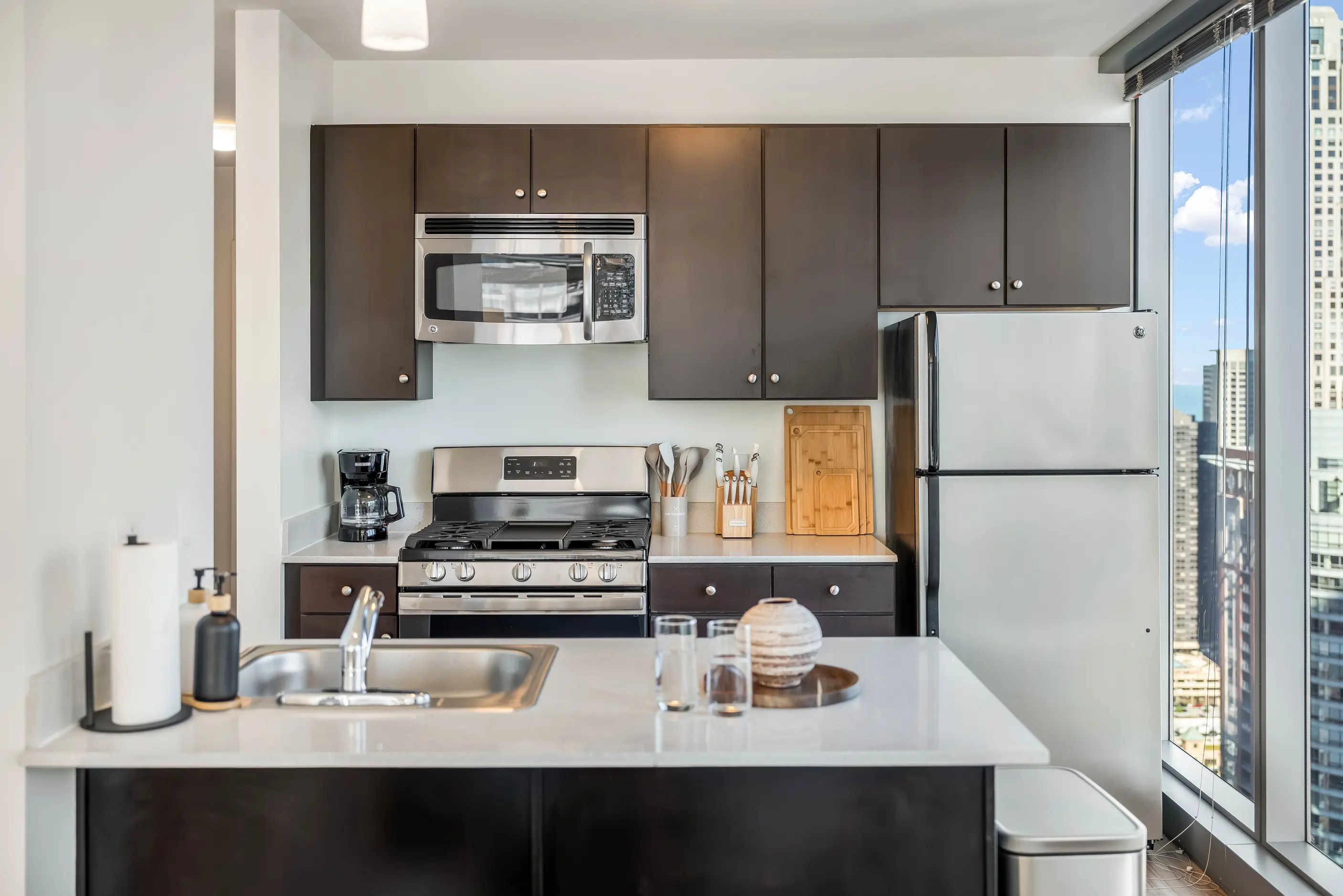 Kitchen breakfast counter with sink beside dark cabinets and tall city-view window at The Atlas Highrise in Chicago