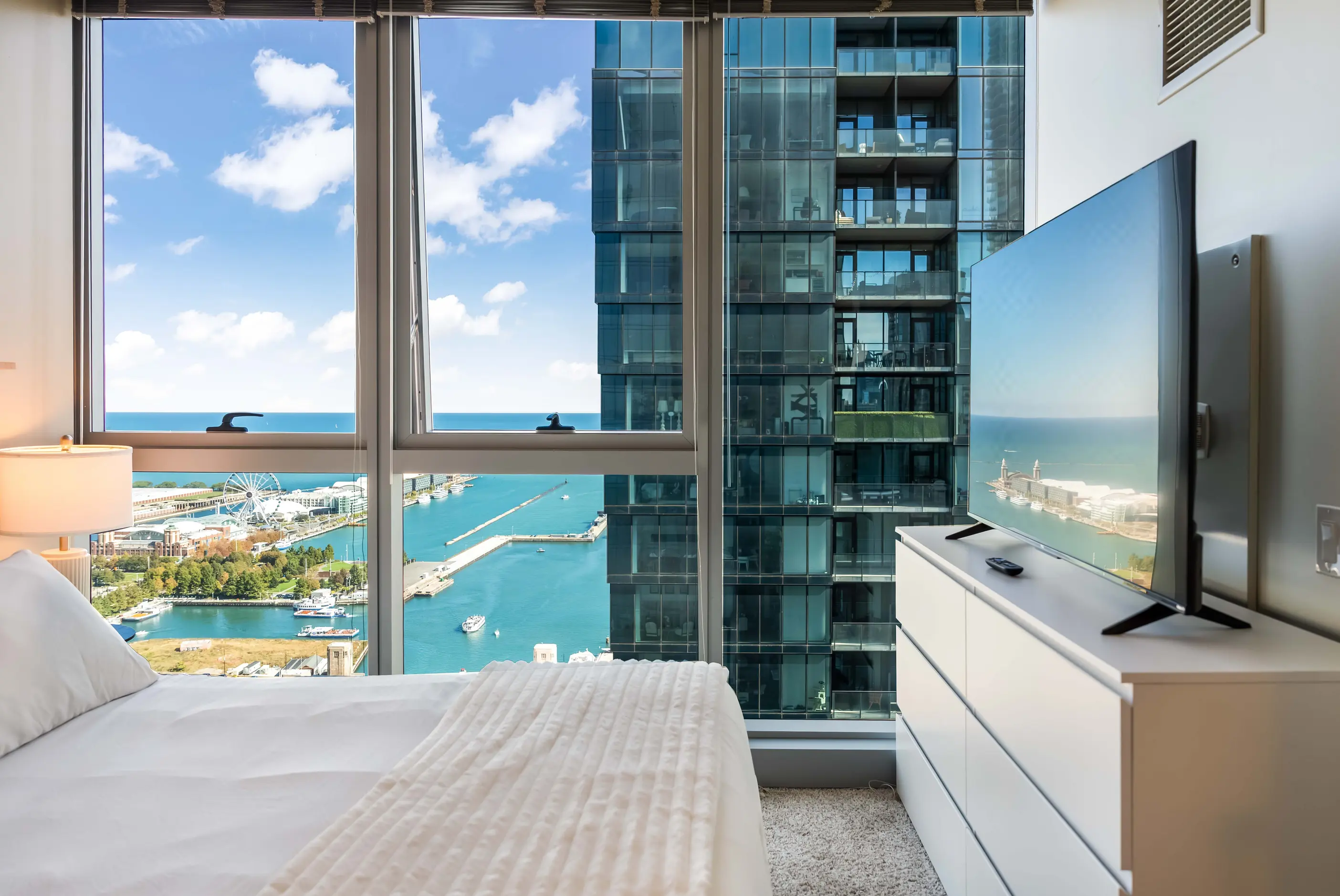 Bedroom with white bedding, white dresser, television, and tall windows overlooking a blue marina at The Atlas Highrise in Chicago