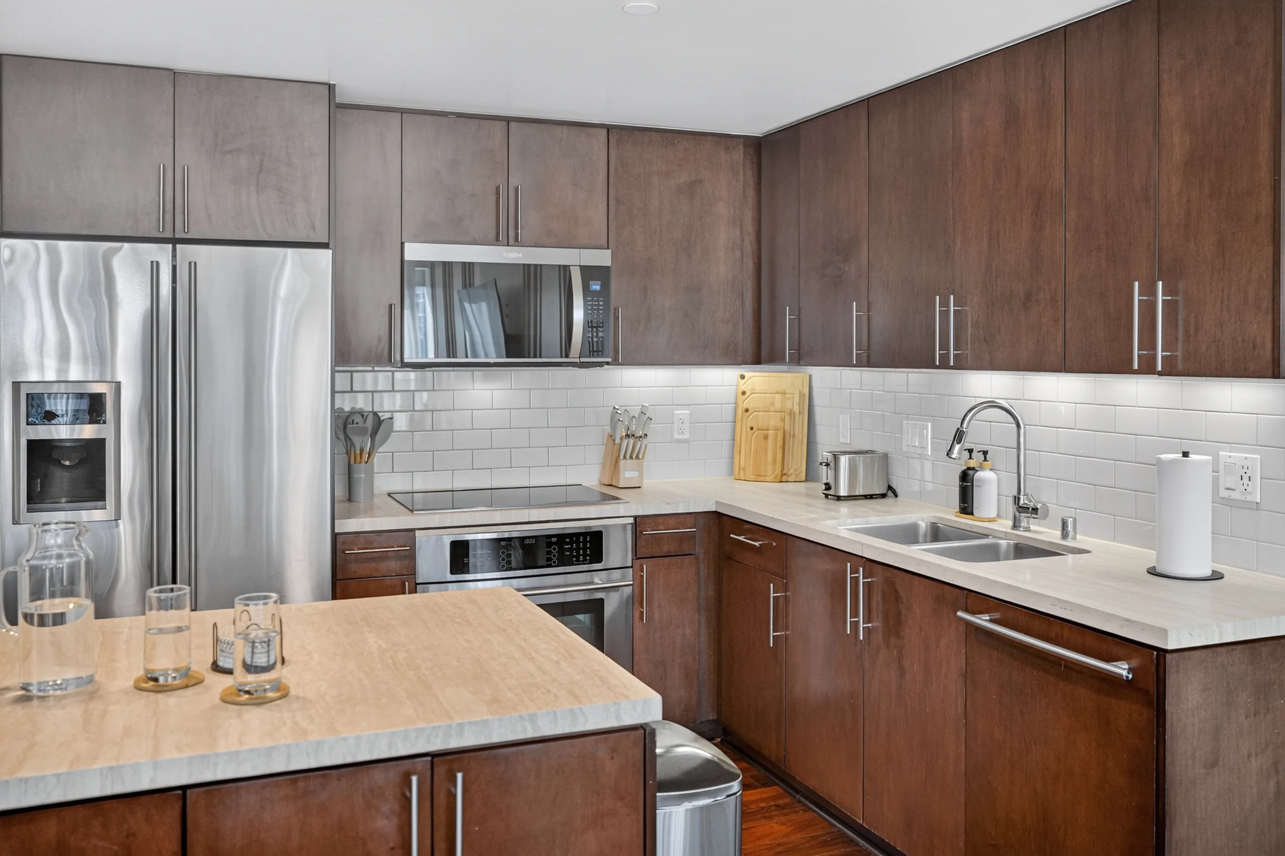 Kitchen with dark cabinets, stainless appliances, white tiled backsplash, and glasses on an island at The Meridian Highrise in Los Angeles