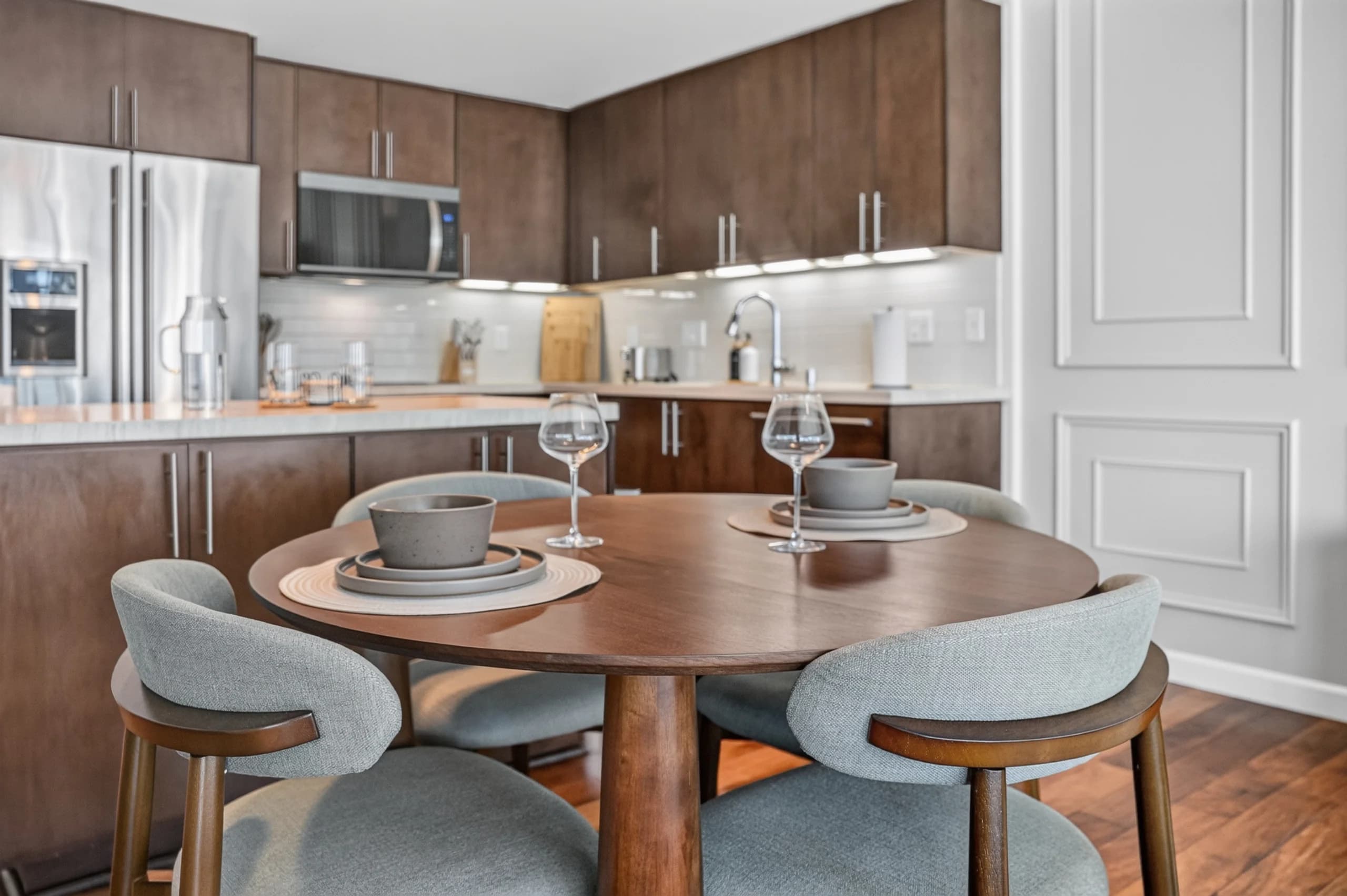 Dining table with cup settings and pale chairs in front of a dark-cabinet kitchen at The Meridian Highrise in Los Angeles