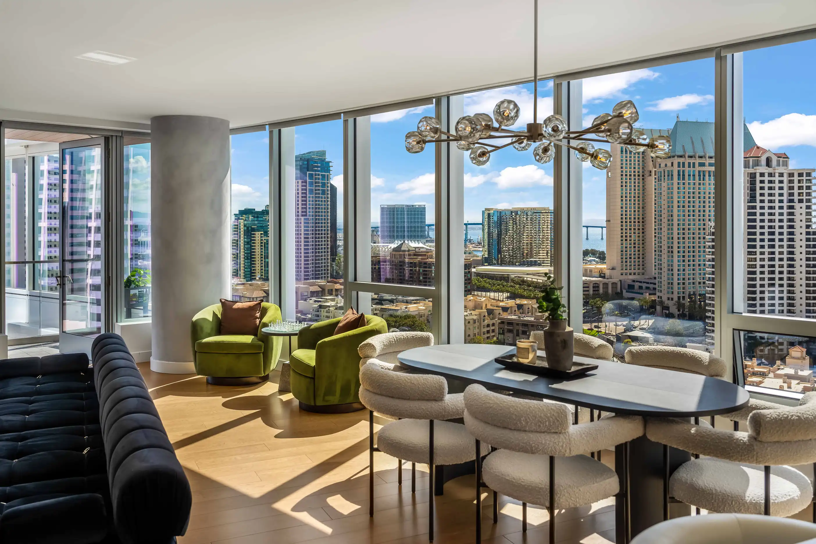 Living and dining area with dark sofa, green chairs, pale seating, chandelier, and full-height windows at Pacific Gate Flagship by World Class® in San Diego