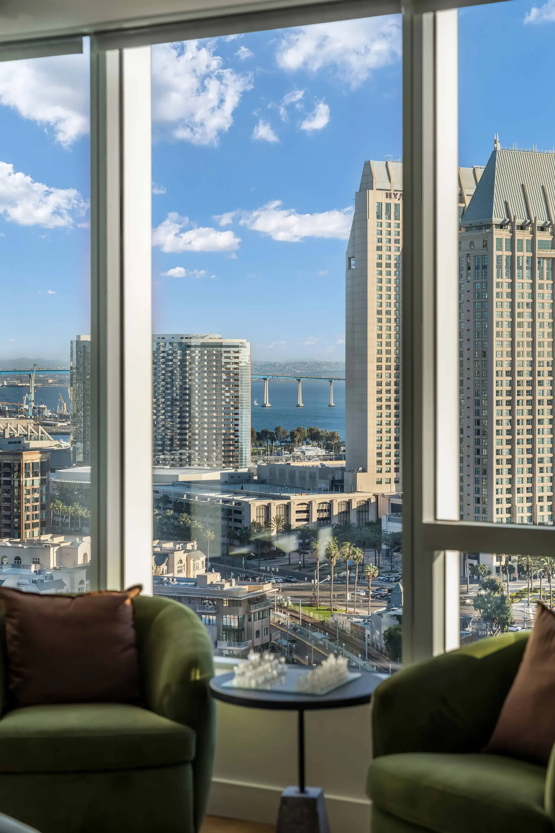 Two green armchairs beside small table facing tall windows overlooking city and distant water at Pacific Gate Flagship in San Diego