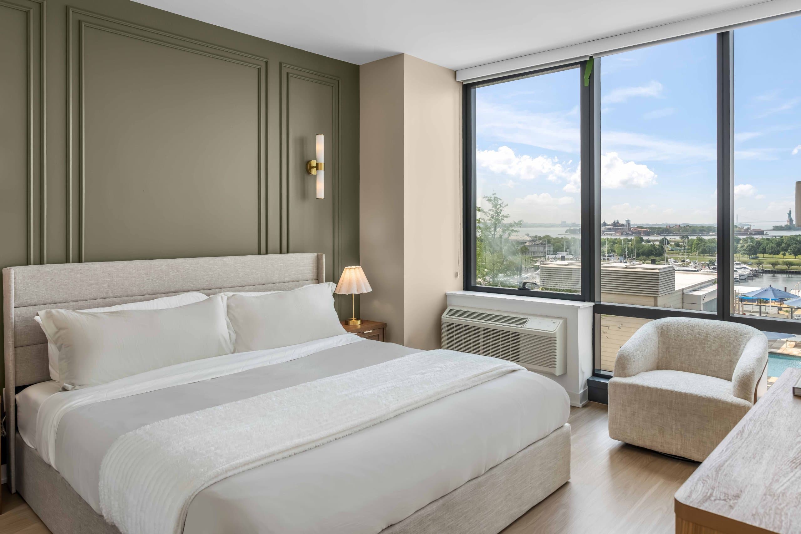 Bedroom with green paneled wall, white bedding, bedside lamp, and armchair beside tall windows at The Metropolis in Jersey City